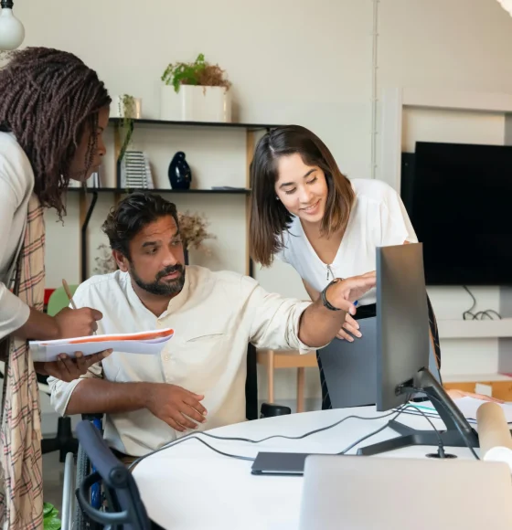 A woman pointing at a computer screen, a man sitting in front of it, and another woman standing beside them.